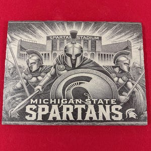 Può includere: Illustrazione in bianco e nero di tre guerrieri spartani in piedi di fronte allo stadio Spartan. Lo stadio ha le parole "Spartan Stadium" sopra l'ingresso. I guerrieri tengono lance e scudi. Il testo "Michigan State Spartans" è sotto i guerrieri.