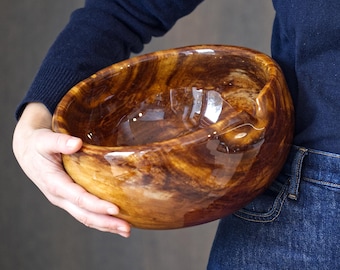 Puede incluir: Un cuenco grande de madera tallada a mano con un acabado brillante. El cuenco tiene un color marrón cálido y rico con patrones de vetas de madera más claros. El cuenco es sostenido por una persona que lleva un suéter azul marino y vaqueros.