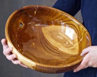Puede incluir: Un cuenco grande y ovalado de madera con un acabado brillante. El cuenco está sostenido por una persona. La veta de la madera es visible, con tonos marrones y beige. Un pequeño agujero es visible cerca del borde superior del cuenco. El cuenco es probablemente para servir o exhibir.