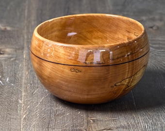 Puede incluir: Un tazón de madera con un acabado de veta de madera natural. El tazón tiene una banda marrón oscura alrededor del borde superior y un acabado brillante.