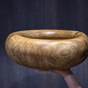 Può includere: Una grande ciotola in legno arrotondata caratterizzata da un bordo spesso e ricurvo verso l'interno. La ciotola presenta una venatura naturale del legno color marrone chiaro con nodi circolari visibili e texture organiche sulla superficie.
