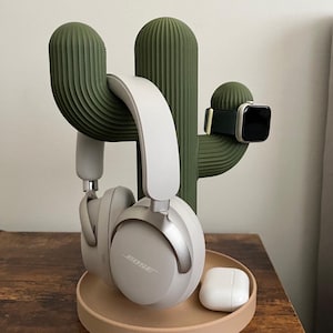 May include: A green cactus-shaped headphone and watch stand. The stand holds a pair of white headphones, a smartwatch with a black screen and a pair of white earbuds. The stand is on a wooden surface.