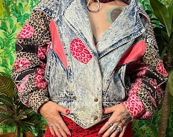 Può includere: Una giacca in denim lavaggio acido con maniche leopardate, toppe leopardate rosa e borchie in metallo argentato sulle spalle. La giacca presenta un ampio colletto, inserti in tessuto rosa e cinturini neri sulle maniche. Sotto, una gonna rossa.