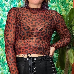 May include: A long-sleeve, mock-neck crop top featuring a sheer, rust-orange fabric with a black leopard print pattern. The top is worn over a black undergarment and paired with black pants that have a front lace-up closure with silver grommets.