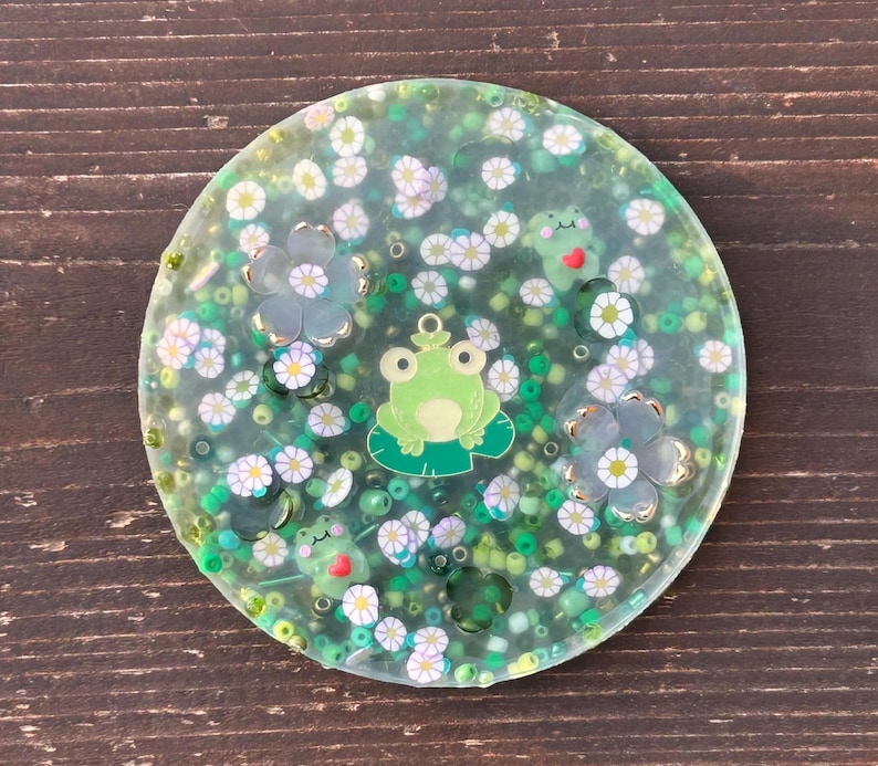 May include: A clear round resin coaster with a green frog, green and white flowers, and green and white beads.