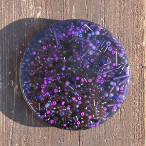 May include: A round, clear resin coaster with embedded purple, pink, and blue glitter and beads.