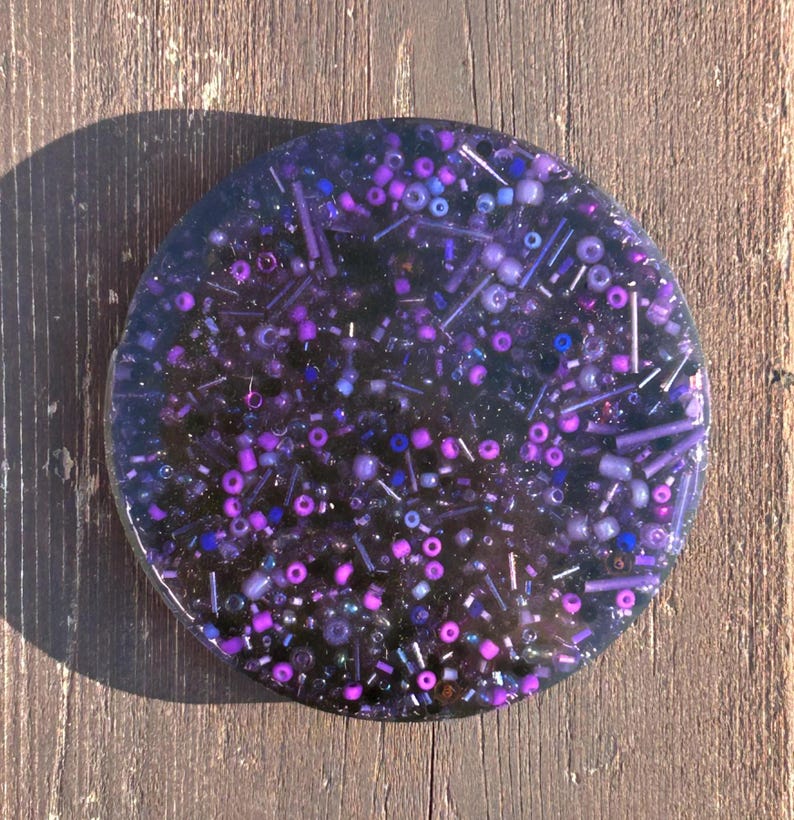 May include: A round, clear resin coaster with embedded purple, pink, and blue glitter and beads.