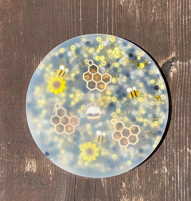 Puede incluir: Un posavasos redondo transparente con un dise&ntilde;o de abeja y panal. El posavasos est&aacute; decorado con cuentas amarillas, negras y blancas.