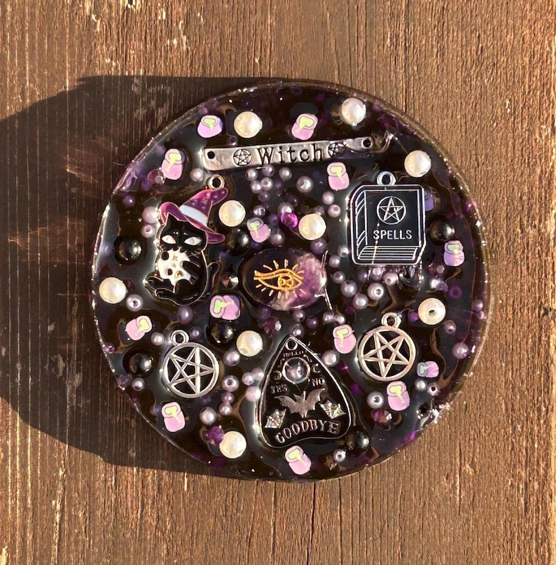 May include: A round, purple resin tray with a black cat, a witch's hat, a book with the word "Spells" on it, a pentagram, a Ouija board, and a crystal eye. The tray is decorated with white pearls and purple glitter.