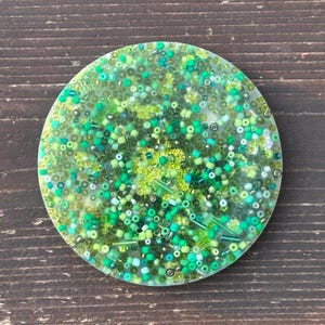 May include: A round, clear resin coaster with a variety of green, blue, and white beads embedded in the resin.