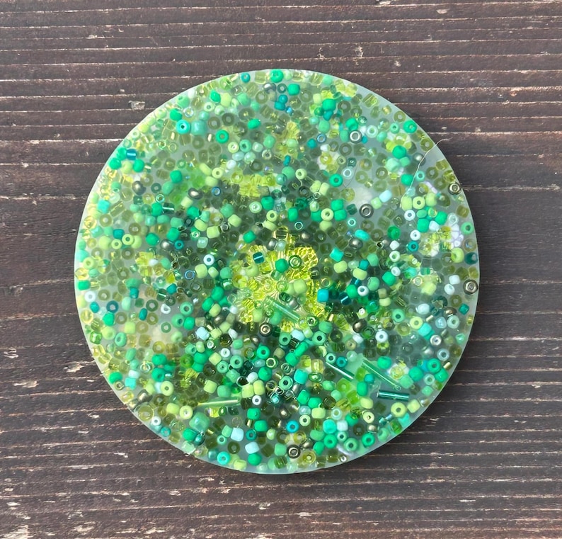 May include: A round, clear resin coaster with a variety of green, blue, and white beads embedded in the resin.