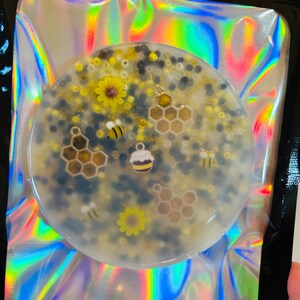 Puede incluir: Un amuleto redondo de resina transparente con tem&aacute;tica de abejas. El amuleto est&aacute; lleno de cuentas negras y blancas, abejas amarillas y negras, amuletos de panal y flores amarillas.