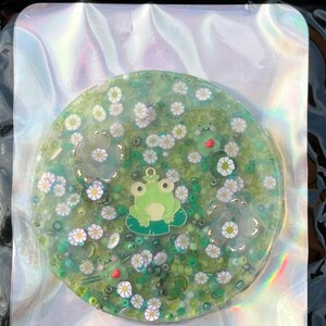 May include: A round, green resin coaster with a frog, white flowers, and green glitter. The coaster is on a holographic background.