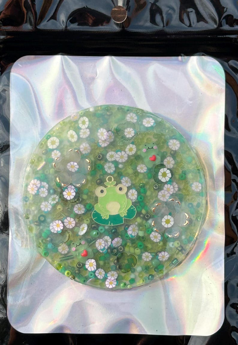 May include: A round, green resin coaster with a frog, white flowers, and green glitter. The coaster is on a holographic background.