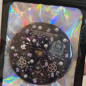 May include: A black resin charm with a witch theme. It features a black cat wearing a witch hat, a pentagram, a book with the word "spells" on it, a Ouija board, and a cauldron. The charm is decorated with silver and white beads.