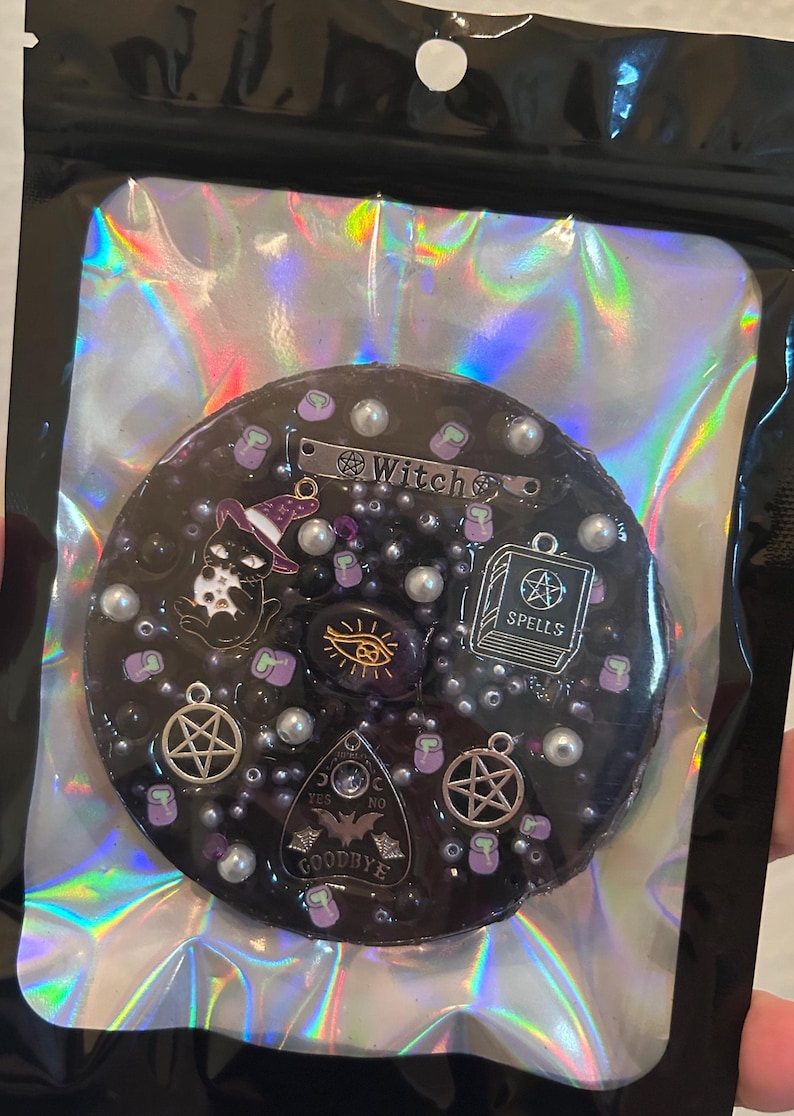 May include: A black resin charm with a witch theme. It features a black cat wearing a witch hat, a pentagram, a book with the word "spells" on it, a Ouija board, and a cauldron. The charm is decorated with silver and white beads.