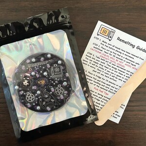 May include: A black and silver resin charm with a witch hat, eye, and other spooky charms. The charm is on a black background with a holographic finish. The image also includes a wooden craft stick and a remelting guide with instructions for how to melt and re-use the charm.