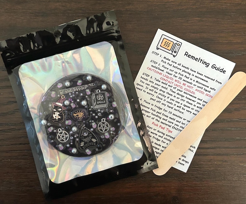 May include: A black and silver resin charm with a witch hat, eye, and other spooky charms. The charm is on a black background with a holographic finish. The image also includes a wooden craft stick and a remelting guide with instructions for how to melt and re-use the charm.