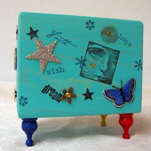 May include: Teal blue wooden box with decorative embellishments, including stars, a butterfly, and a woman's face. The box has the words "faith", "hope", "wish", and "dream" written on it. The box is sitting on three legs, one blue, one red, and one yellow.