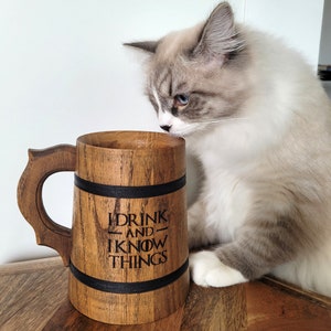 May include: A wooden beer stein with a black band and the text "I drink and I know things" engraved on the side. A cat is sniffing the stein.
