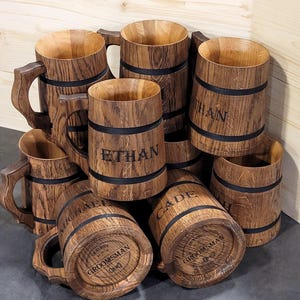 Puede incluir: Un grupo de ocho jarras de cerveza de madera con bandas negras y nombres personalizados. Las jarras están apiladas unas sobre otras y tienen un aspecto rústico. Una jarra tiene el texto "Groomsman" grabado.