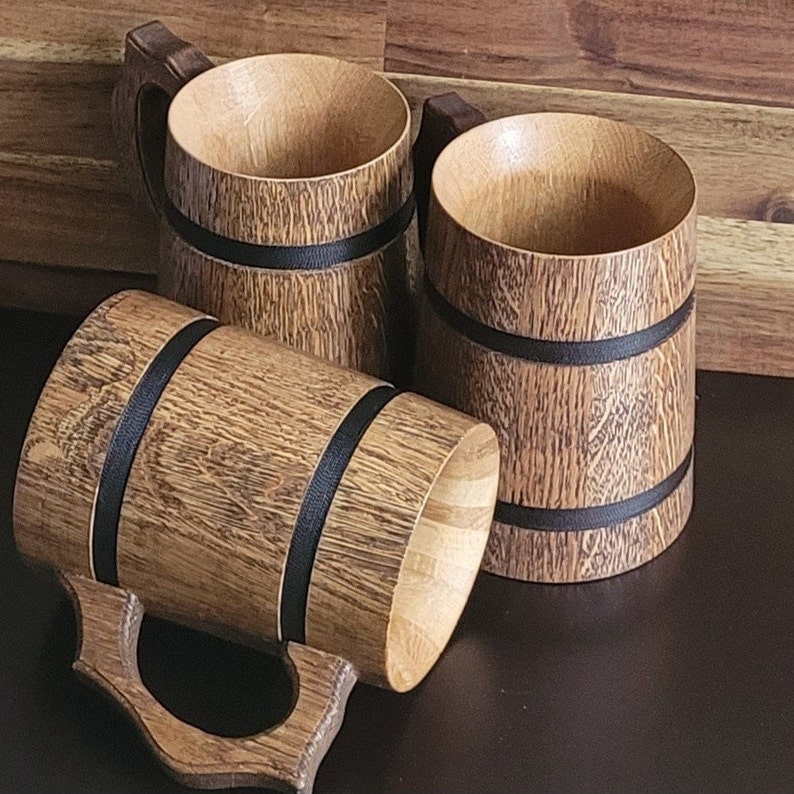 May include: Three wooden beer mugs with black bands around the top. The mugs are made of a light brown wood with a natural grain pattern.