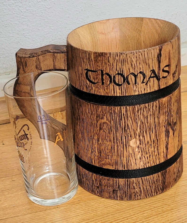 May include: A wooden beer stein with a black band around the top and bottom. The stein is engraved with the name "Thomas".