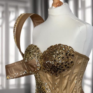 Puede incluir: Un vestido dorado sin tirantes con un corpiño de corsé y una tira de un solo hombro. El vestido está cubierto de brillantes pedrerías y cuentas.