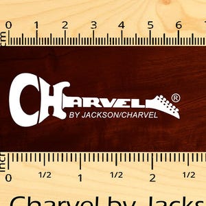 May include: A waterslide decal for a Charvel by Jackson guitar. The decal is solid white and measures 5 inches long. The decal is shown on a ruler with inch markings.