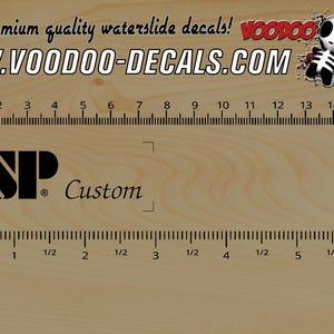 Może przedstawiać: Czarno-białe logo ESP Custom z tekstem "ESP Custom" i symbolem znaku towarowego. Logo znajduje się na drewnianej powierzchni z linijką w centymetrach i calach.