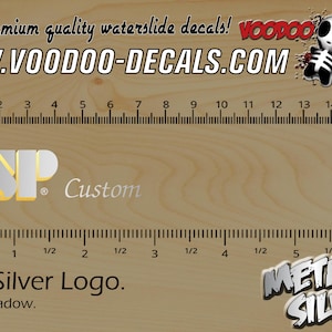 May include: A waterslide decal featuring the ESP logo in silver with a gold shadow. The decal is 2 inches wide and is part of a set of high-quality waterslide decals. The decal is shown on a wooden surface with a ruler for scale.