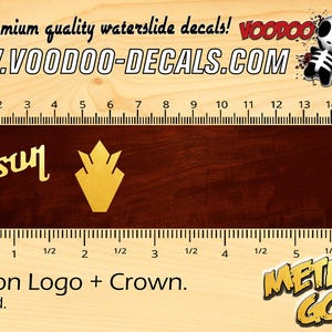 May include: Waterslide decal sheet featuring a gold Gibson logo and crown. The decal sheet is 5 inches long and is on a wooden background. The text "WWW.VOODOO-DECALS.COM" is printed at the top of the image.