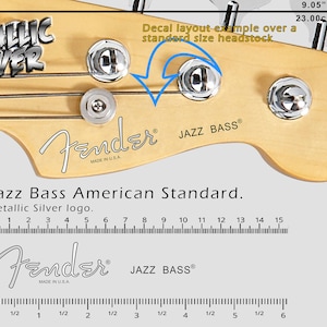 May include: A close-up of a Fender Jazz Bass guitar headstock with a metallic silver decal. The decal reads "Fender" and "JAZZ BASS" with "MADE IN U.S.A." underneath. The headstock is made of natural wood and has four tuning pegs. A ruler is shown below the headstock with measurements in inches and centimeters.