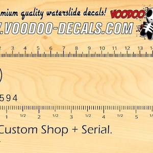 Könnte beinhalten: Ein Lineal mit Markierungen in Zoll und Zentimetern, das ein Wasserschiebebild für ein Skateboard mit dem Text "ESP Custom Shop + Serial. All Black." und der Website "WWW.VOODOO-DECALS.COM" zeigt.