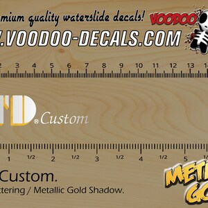 Könnte beinhalten: Holzhintergrund mit einem Lineal in Zoll und Zentimetern. Das Bild zeigt den Text "WWW.VOODOO-DECALS.COM" und "LTD Custom" mit silbernen Buchstaben und einem metallisch goldenen Schatten. Ein Cartoon-Schädel ist ebenfalls vorhanden.