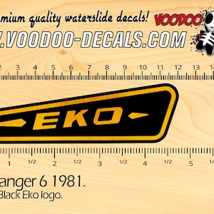 Puede incluir: Calcomanía con el logotipo Eko Ranger 6 1981 en negro y dorado. La calcomanía es rectangular con fondo negro y letras doradas. El texto "EKO" es visible. La imagen también incluye una regla con pulgadas y centímetros.