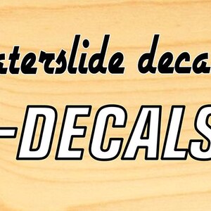 May include: A banner on a light brown wooden background. The text reads "Premium quality waterslide decals!" in a black script font. Below, the website address "WWW.VOODOO-DECALS.COM" is displayed in a bold, white font. A cartoon skeleton character is on the right.