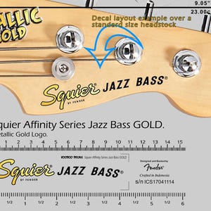 Op de afbeelding: Close-up van de kop van een basgitaar met de tekst "METALLIC GOLD" en "Squier JAZZ BASS". De kop is lichtbruin met zilveren stemknoppen. De breedte is 23,00 cm.
