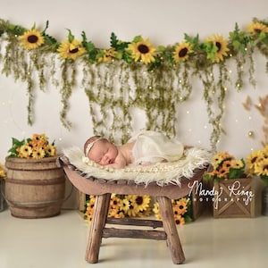 Puede incluir: Un bebé recién nacido yace plácidamente sobre un taburete de madera rústica, envuelto en una suave tela color crema. La escena está decorada con girasoles, vegetación y luces de hadas, creando un ambiente cálido y natural para una sesión de fotos.