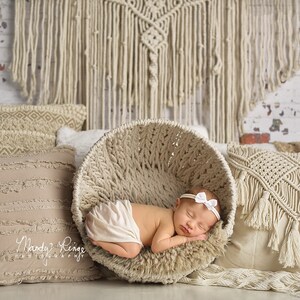 May include: A newborn baby girl sleeping in a woven basket with a white headband. The basket is surrounded by pillows with a neutral color scheme and a macrame wall hanging in the background.