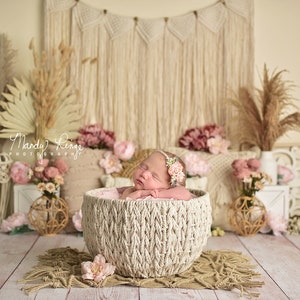 Op de afbeelding: Een pasgeboren babymeisje slaapt in een geweven mand die versierd is met roze bloemen. De mand is omgeven door een bohemian achtergrond met macramé wandhangers, pampasgras en andere natuurlijke elementen.