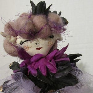 May include: A handmade doll with a purple and black dress, a pink wig, and a black crown. The doll has a white face with embroidered features and pink cheeks.