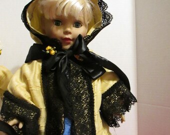 May include: A doll wearing a yellow and black hooded jacket with lace trim. The doll has blonde hair and blue eyes.