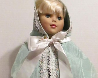 May include: A doll wearing a light green hooded cape with white lace trim and a white satin bow. The cape has a delicate floral pattern and the doll has blonde hair.