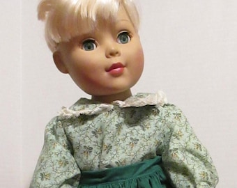 Könnte beinhalten: Eine Puppe mit blonden Haaren, die ein grünes Kleid mit einem weißen Spitzenkragen und einer Bluse mit Blumenmuster trägt.