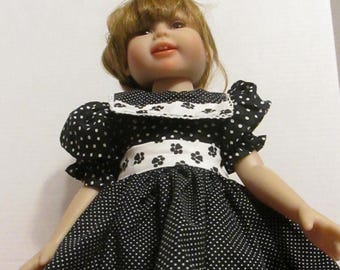 May include: A black and white polka dot dress with a white collar and a white belt with black paw prints. The dress is being worn by a doll with brown hair and a white face.
