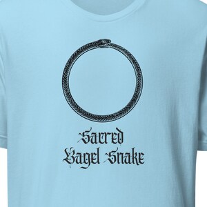 May include: Light blue t-shirt featuring a black ouroboros design, a circular symbol of a snake eating its own tail. The text "Sacred Bagel Snake" is printed below the ouroboros in a gothic font. The shirt is made of soft material.