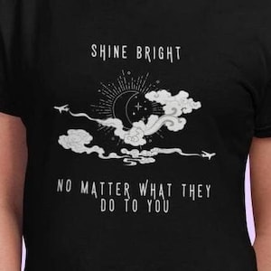 Puede incluir: Camiseta negra con un diseño gráfico blanco de una luna, estrellas y nubes. El texto "Shine Bright No Matter What They Do To You" está impreso en la camiseta.