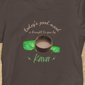 以下が含まれることがあります： 茶色のTシャツに「today's good mood is brought to you by... Kava」という文字と、茶色の液体と緑の葉が入った木製のボウルを描いたグラフィックがプリントされています。