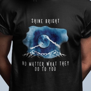 Puede incluir: Camiseta negra con un diseño de acuarela de una luna, montañas y nubes. El texto "Shine Bright No Matter What They Do To You" está impreso en la camiseta.